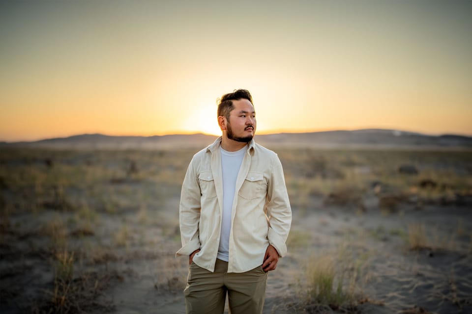 Man in a cream shirt standing in a desert field at golden hour, the setting sun on the horizon behind him