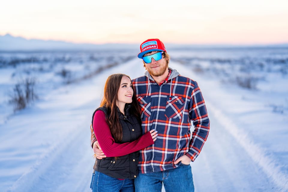 Couple standing together on a snowy road at dusk, the man in a red cap, mirrored sunglasses and a plaid shirt