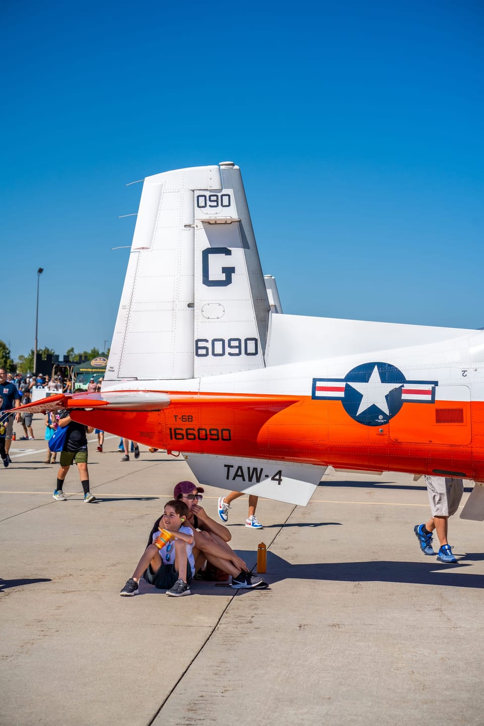 White and orange Navy trainer aircraft on the tarmac at an airshow, with spectators resting in the tail's shade