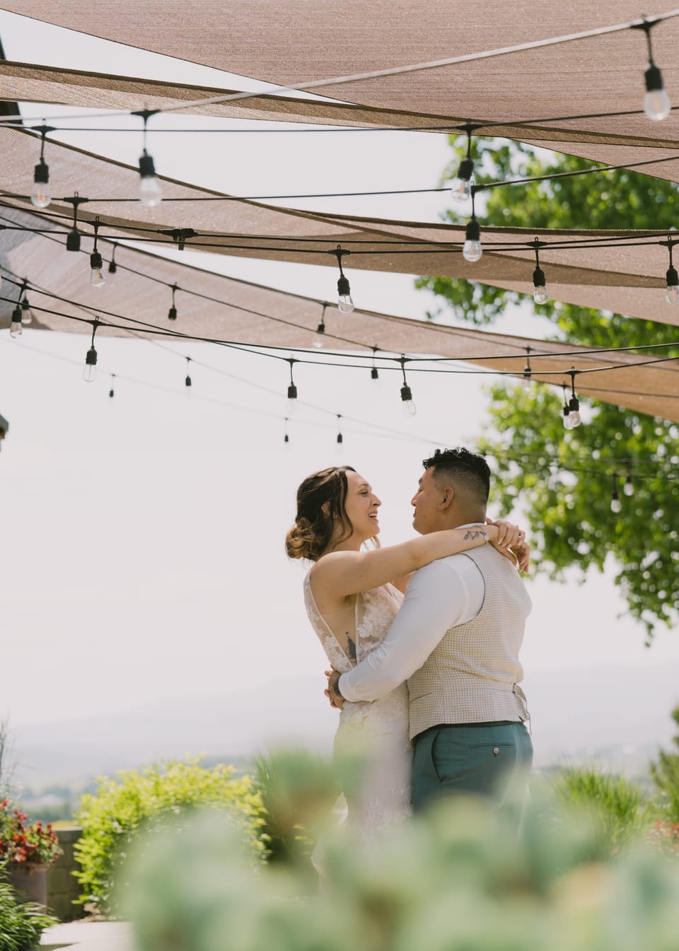 Bride and groom dancing under a canopy of string lights, greenery in the foreground