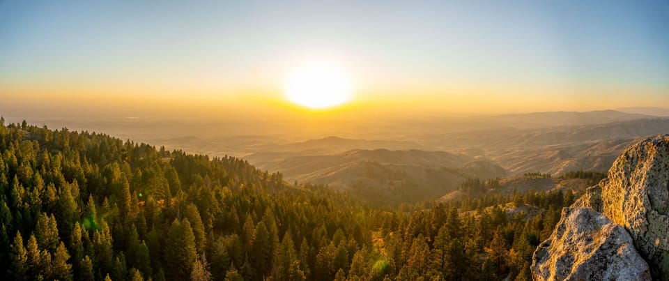 Sun setting over forested mountain ridges, with a rocky outcrop in the right foreground