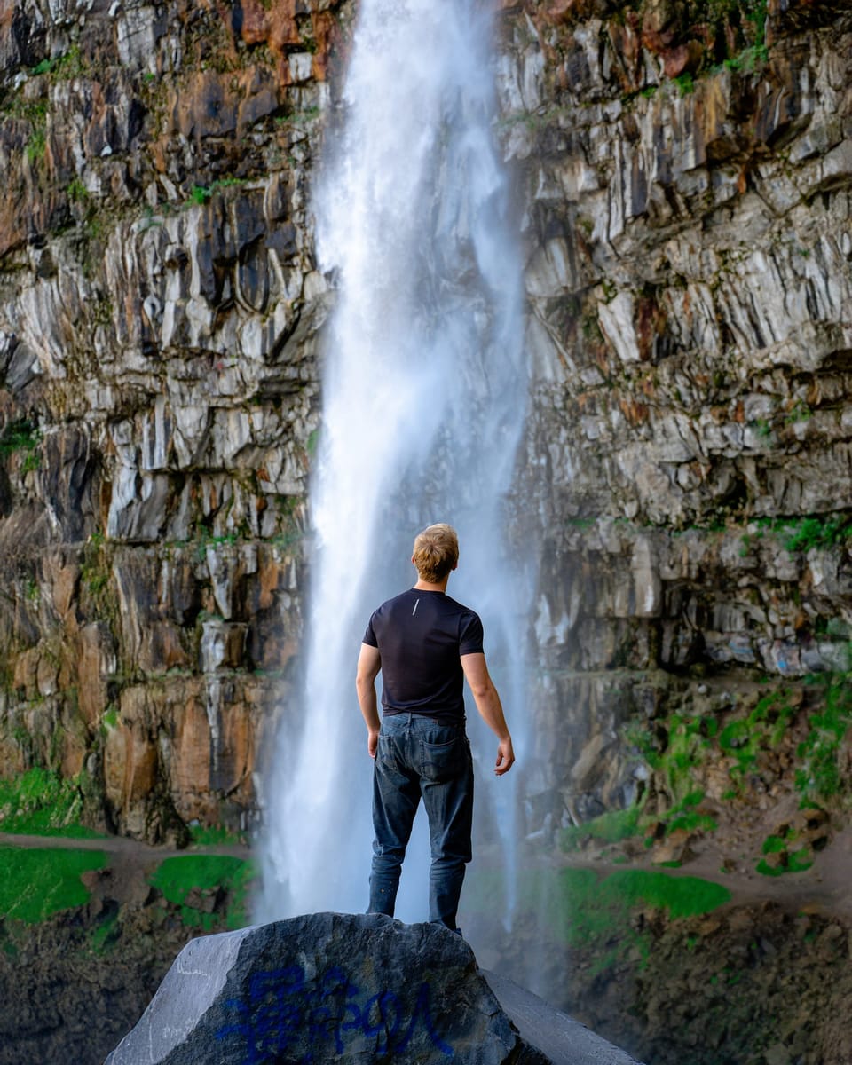Man standing on a boulder at the base of a tall waterfall on a basalt cliff