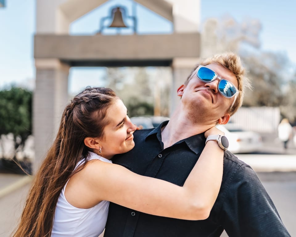 Woman hugging a man in blue mirrored sunglasses in front of a bell tower