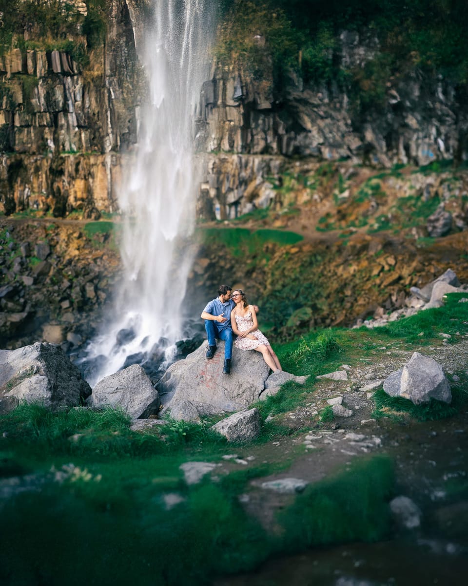 Couple seated on rocks at the base of a tall waterfall in a mossy basalt canyon