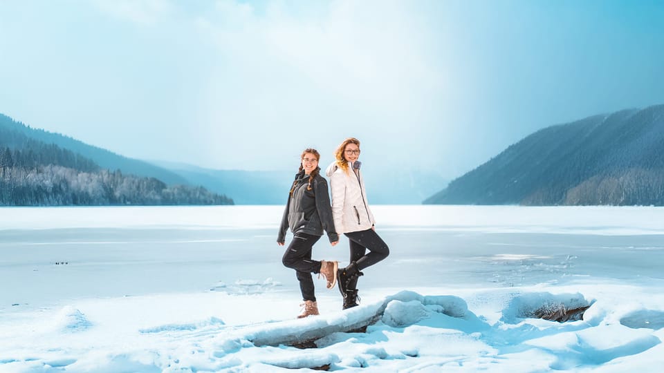 Couple in winter jackets posing on an icy lakeshore with misty blue mountains behind