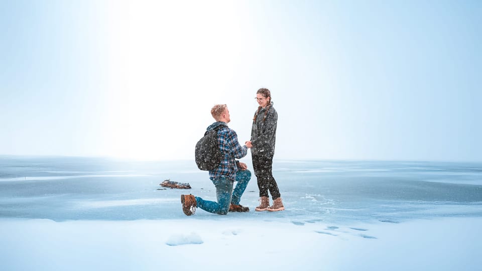 Man kneeling to propose to a woman on a snow-covered frozen lake with pale sky