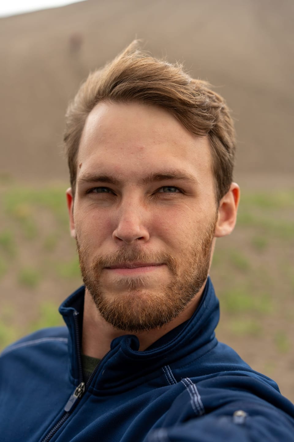 Close headshot of a bearded young man in a navy jacket against a soft green and tan background