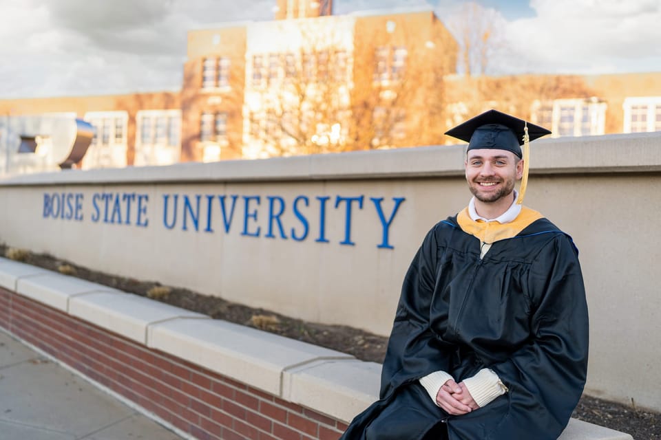 Graduate in cap and gown seated on the Boise State University sign with a campus building behind