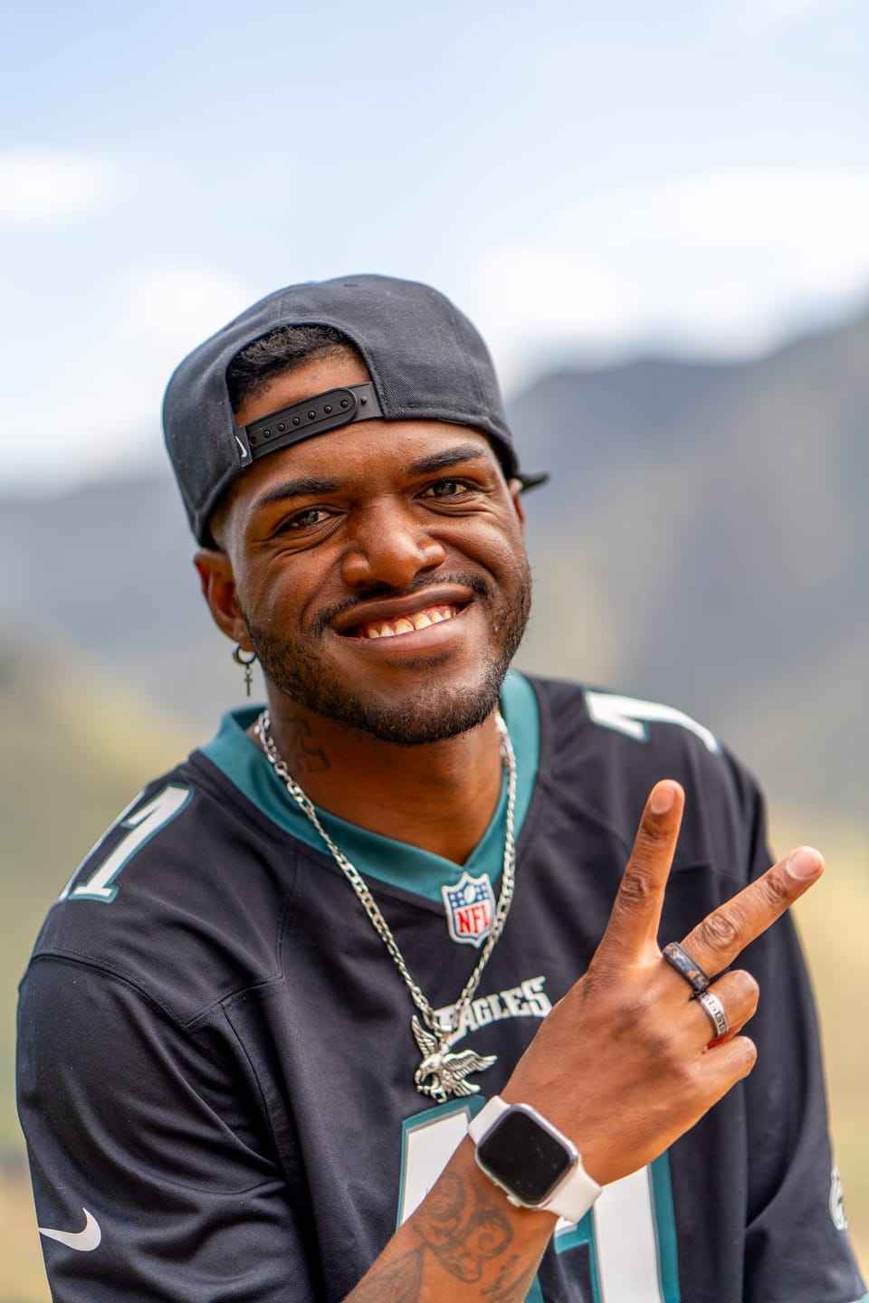 Smiling man in a backwards cap and football jersey making a peace sign, mountains behind him