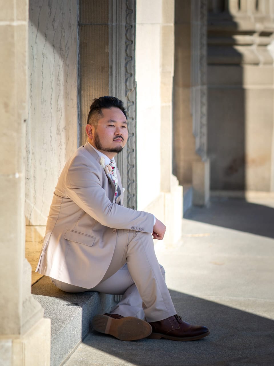 Man in a cream suit seated on stone steps between sunlit columns