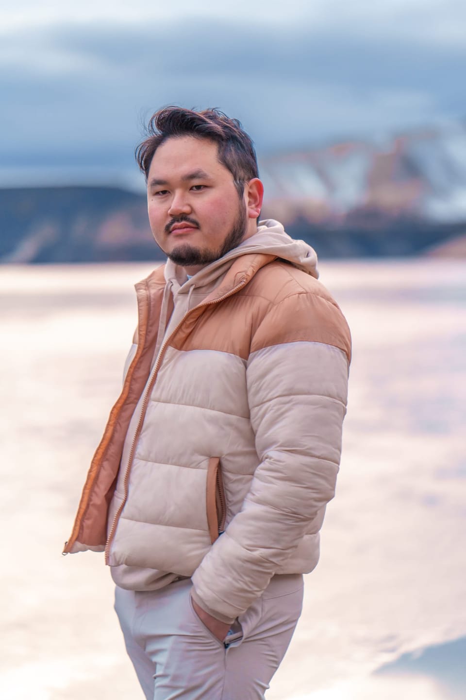 Man in a pink and tan puffer jacket standing in front of a frozen lake and snowy mountains