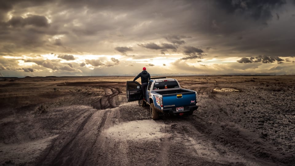 Person standing beside a blue pickup truck on a muddy overlook under dramatic storm clouds and sunbeams