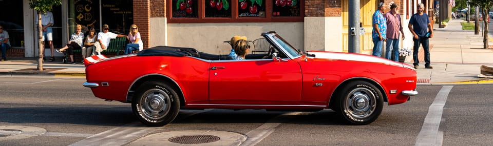 Red first-generation Camaro convertible driving past storefronts on a downtown street