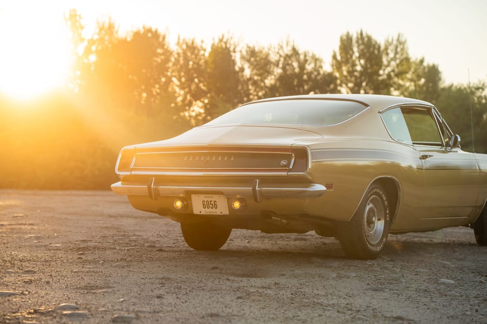Gold classic Plymouth Barracuda parked on gravel with a strong sunset flare through the trees