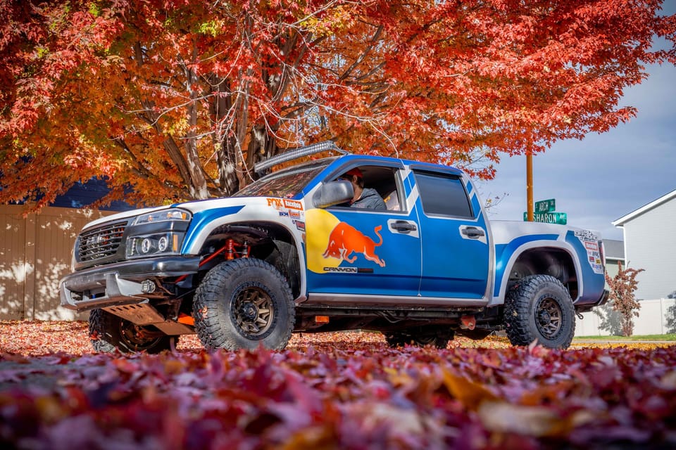 Blue off-road truck with Red Bull livery parked under a bright red autumn tree on fallen leaves