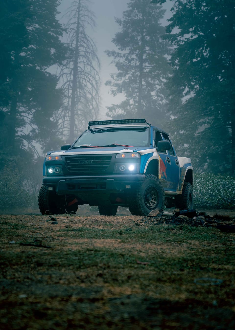 Lifted pickup with glowing headlights and a light bar in a foggy pine forest