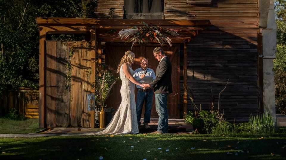 Bride and groom holding hands with their officiant in front of a rustic wooden barn altar decorated with dried florals