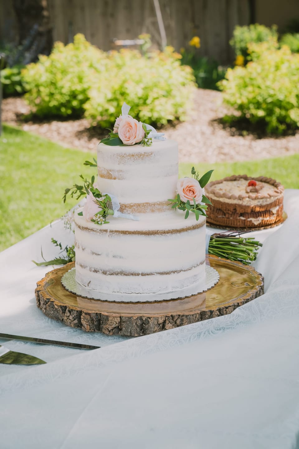 Two-tier semi-naked wedding cake with pink roses on a wood-slice stand outdoors